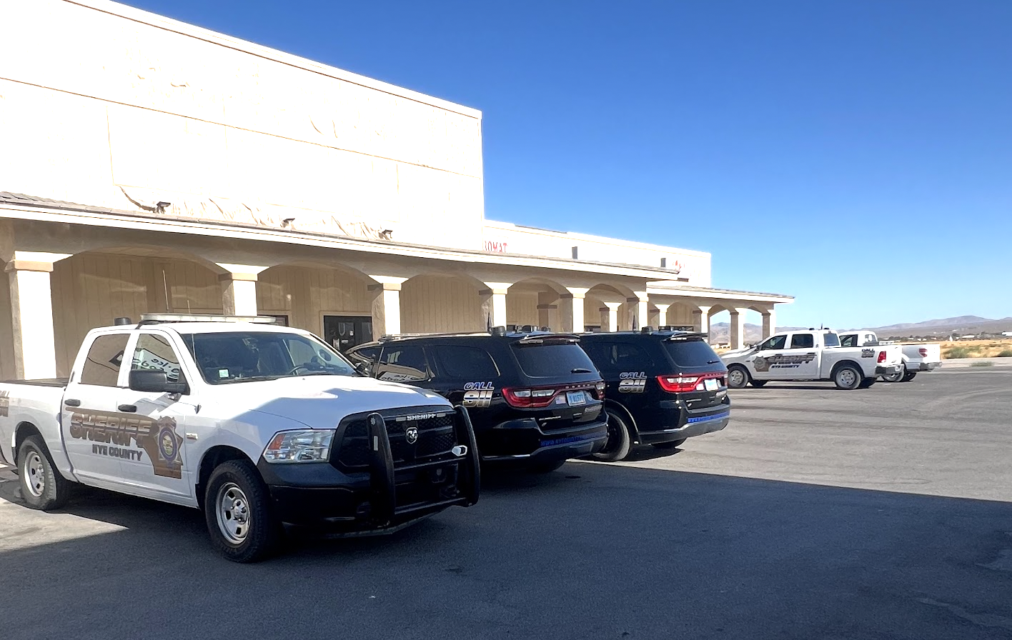 Four Nye County Sheriff vehicles in a parking lot on North Blagg Road in Pahrump. A white Sheriff pickup sits nose out in the foreground, two black marked SUVs are parked nose in to the building beside it, and a second white Sheriff pickup stands further out in the lot. No emergency lights are lit and nobody is in any of them.