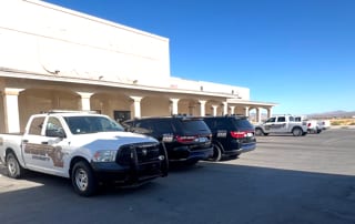 Four Nye County Sheriff vehicles in a parking lot on North Blagg Road in Pahrump. A white Sheriff pickup sits nose out in the foreground, two black marked SUVs are parked nose in to the building beside it, and a second white Sheriff pickup stands further out in the lot. No emergency lights are lit and nobody is in any of them.