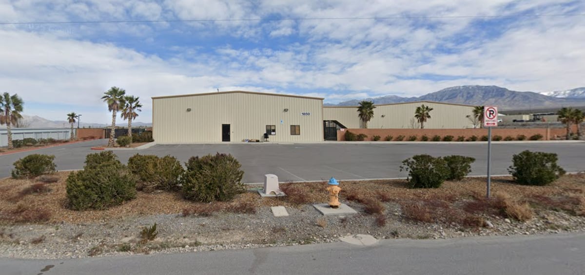 Street view of 1850 East Basin Avenue in Pahrump. A long tan metal building with the number 1850 on it sits behind an empty asphalt lot, with a second tan metal building running east behind a low brick wall. Palm trees, desert scrub and a fire hydrant are in the foreground. Snow-topped mountains are in the distance.
